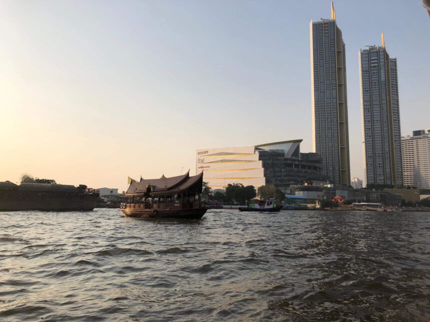 Tajlandia przewodnik -Tradycyjna łódź na rzece Chao Phraya z panoramą nowoczesnych wieżowców Bangkoku o zachodzie słońca.